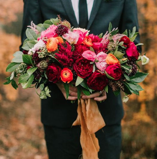 A loose asymmetrical bouquet of Darcey garden roses, Juliet garden roses, Romantic Antique garden roses, burgundy scabiosa, light pink astilbe, peach ranunculus, pink pepper berry, jasmine vine, bay laurel, and seeded eucalyptus wrapped in gold glitter ribbon tied in a bow. 